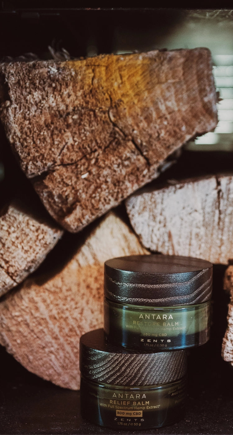 Two jars of Antara Relief Balm and Restore Balm stacked on a dark surface in front of stacked firewood logs.