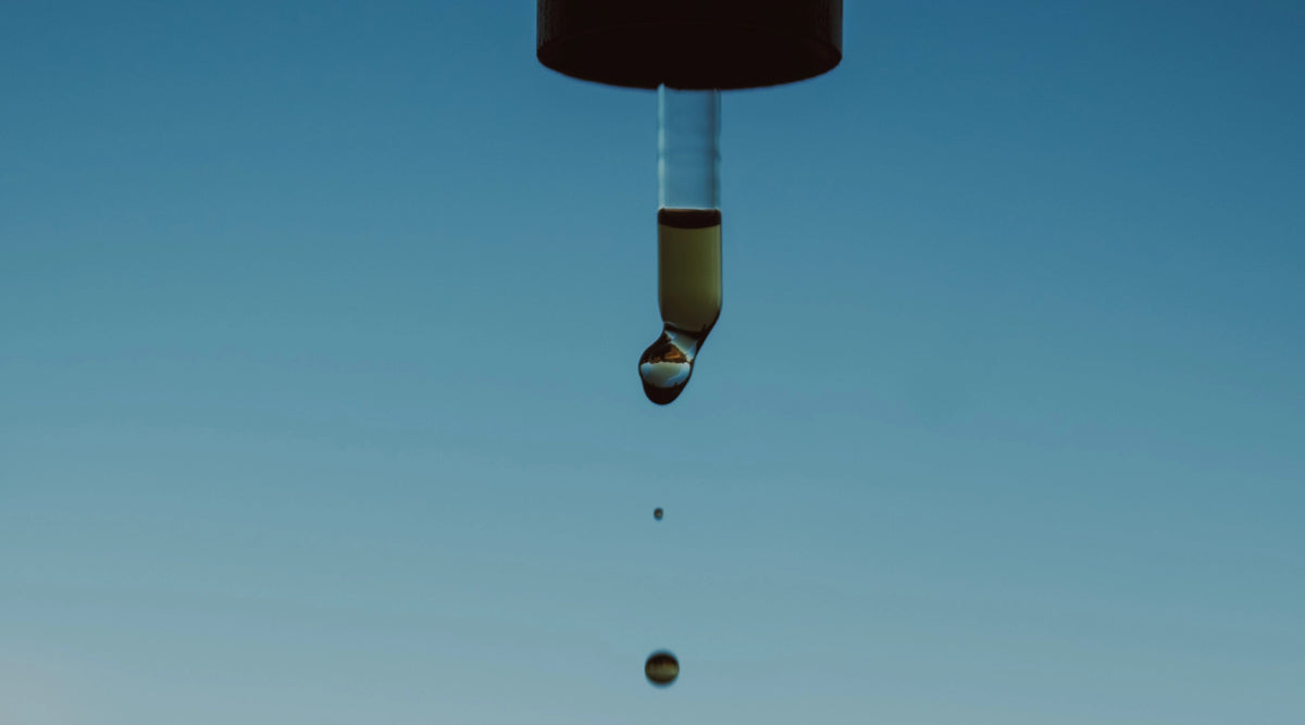 A dropper of Vitality Elixir dripping into the jar with a bright blue natural blurred background.